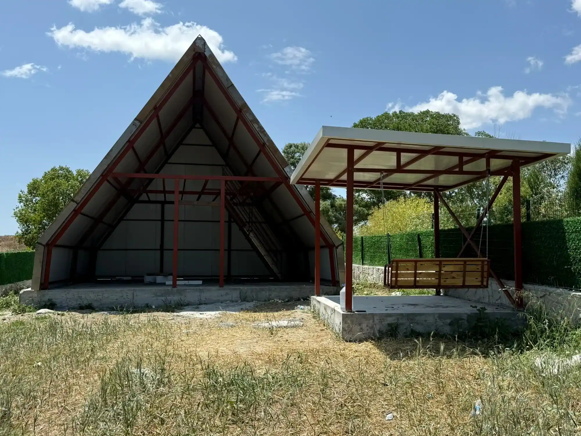 Kırmızı çelik iskeletli A tipi yapı ve yanında salıncaklı çelik pergola